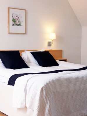 A neatly made bed with white and navy bedding, a wooden headboard, and a wall-mounted lamp. - Kockska Gården (Simrishamn)