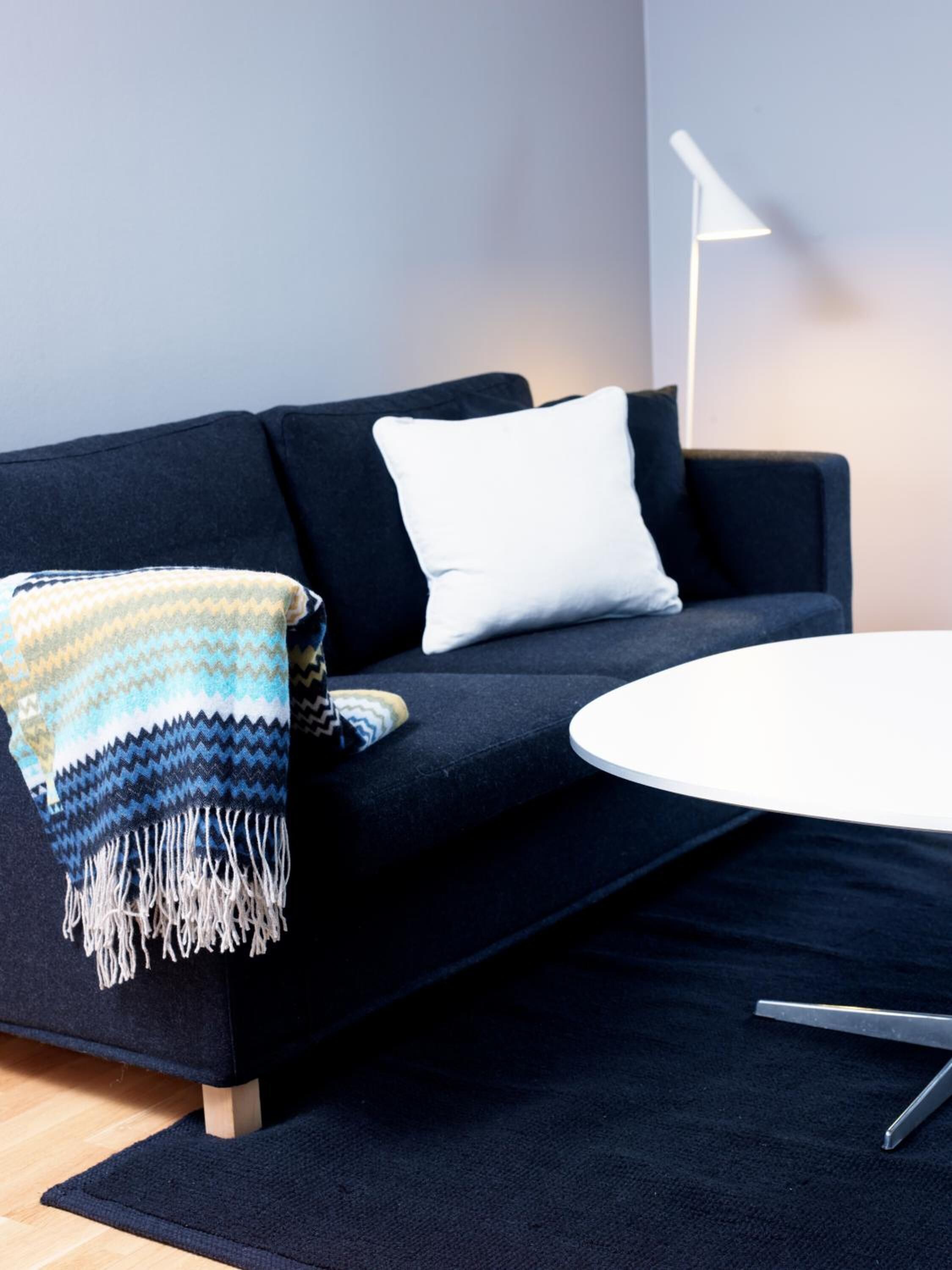A modern living room with a dark sofa, a white pillow, a patterned throw blanket, a round white table, and a floor lamp.