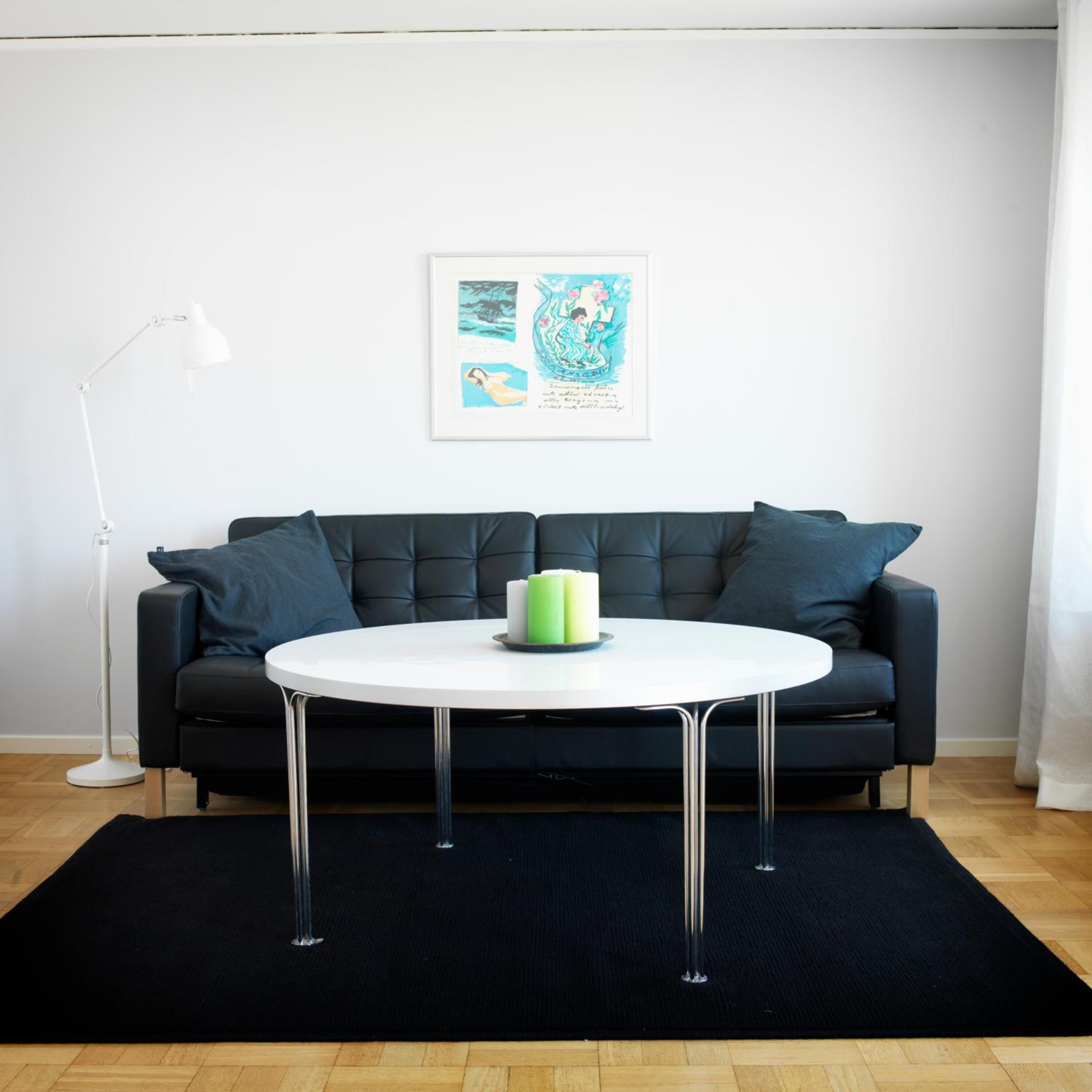 Una sala de estar moderna con un sofá negro, una mesa redonda blanca con velas verdes, y una lámpara de pie.