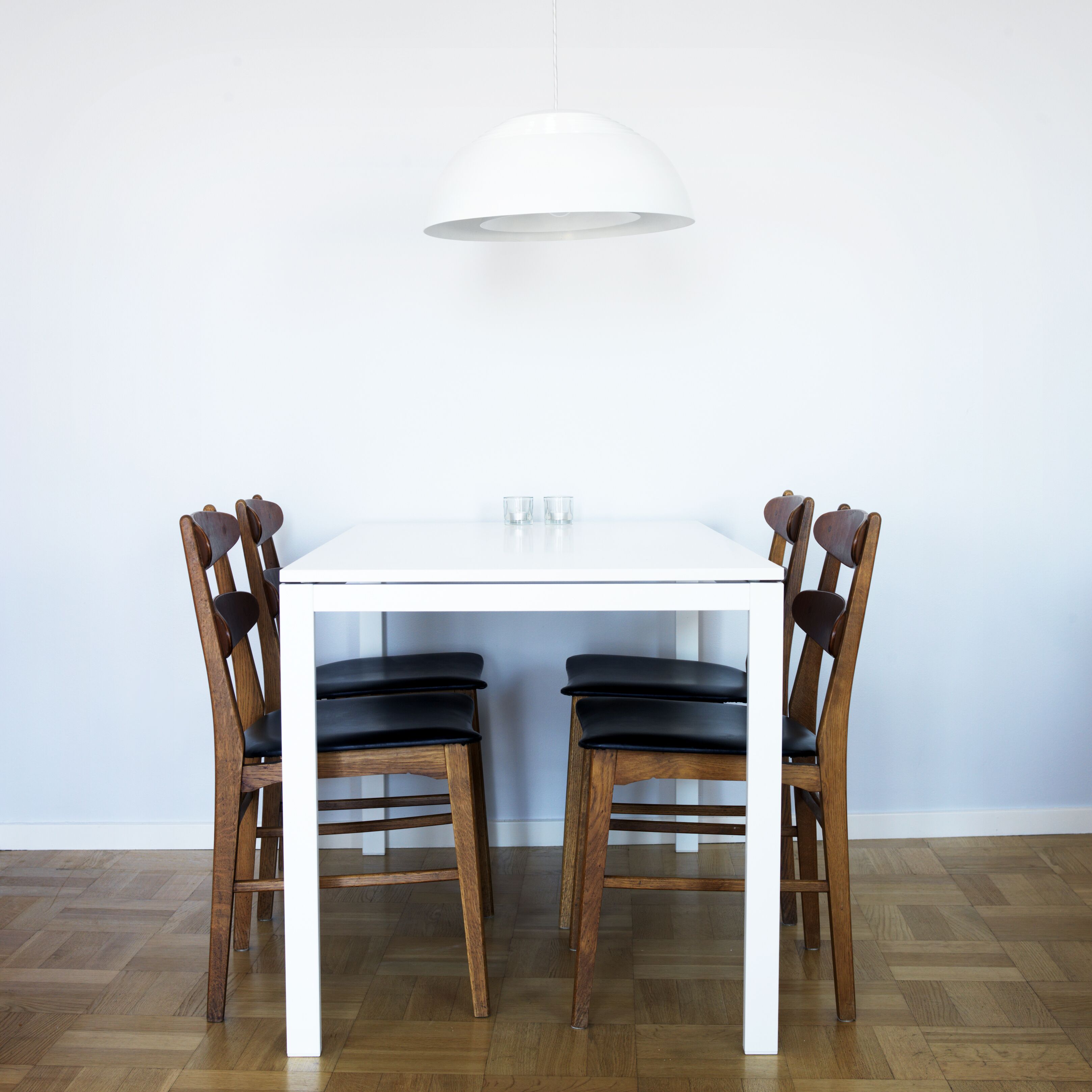 Una mesa de comedor blanca con cuatro sillas de madera, una lámpara colgante blanca y una pared blanca.