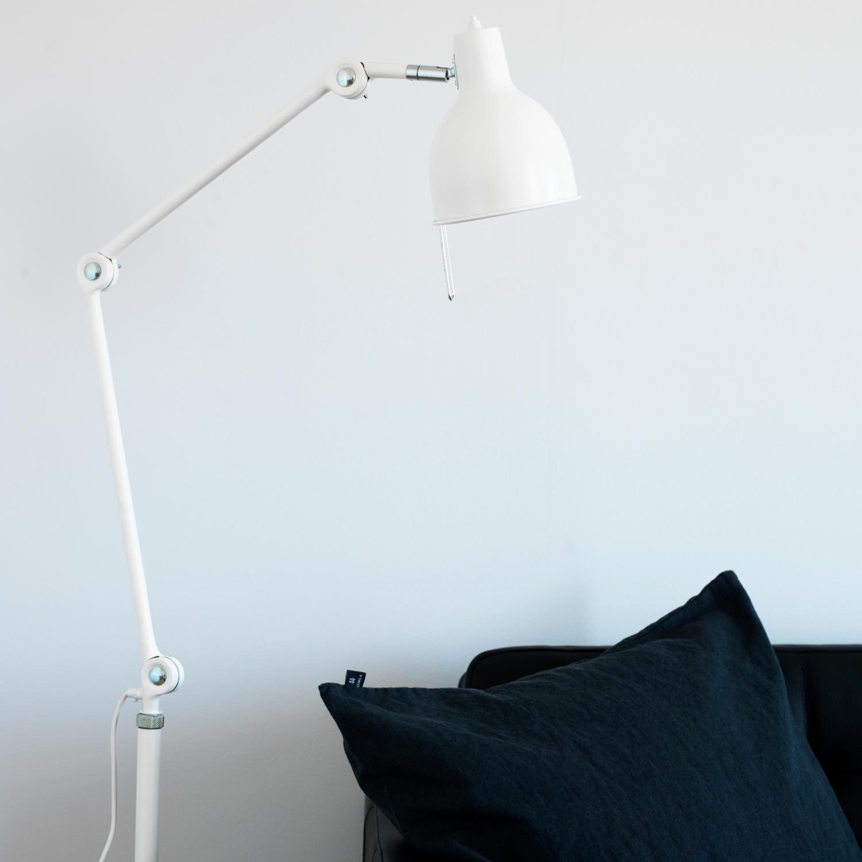 Une lampe de bureau blanche, dotée d’un bras flexible et d’un abat-jour blanc, est placée à côté d’un coussin bleu foncé sur une chaise noire, le tout adossé à un mur uni.