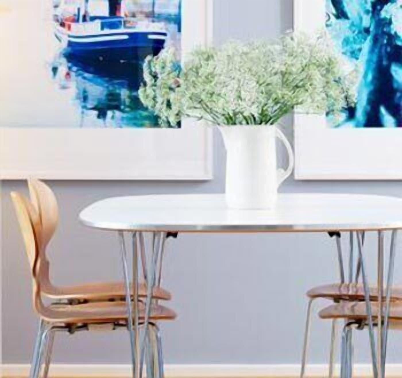 A dining table with a white pitcher of flowers, two wooden chairs, and framed artwork on the wall.