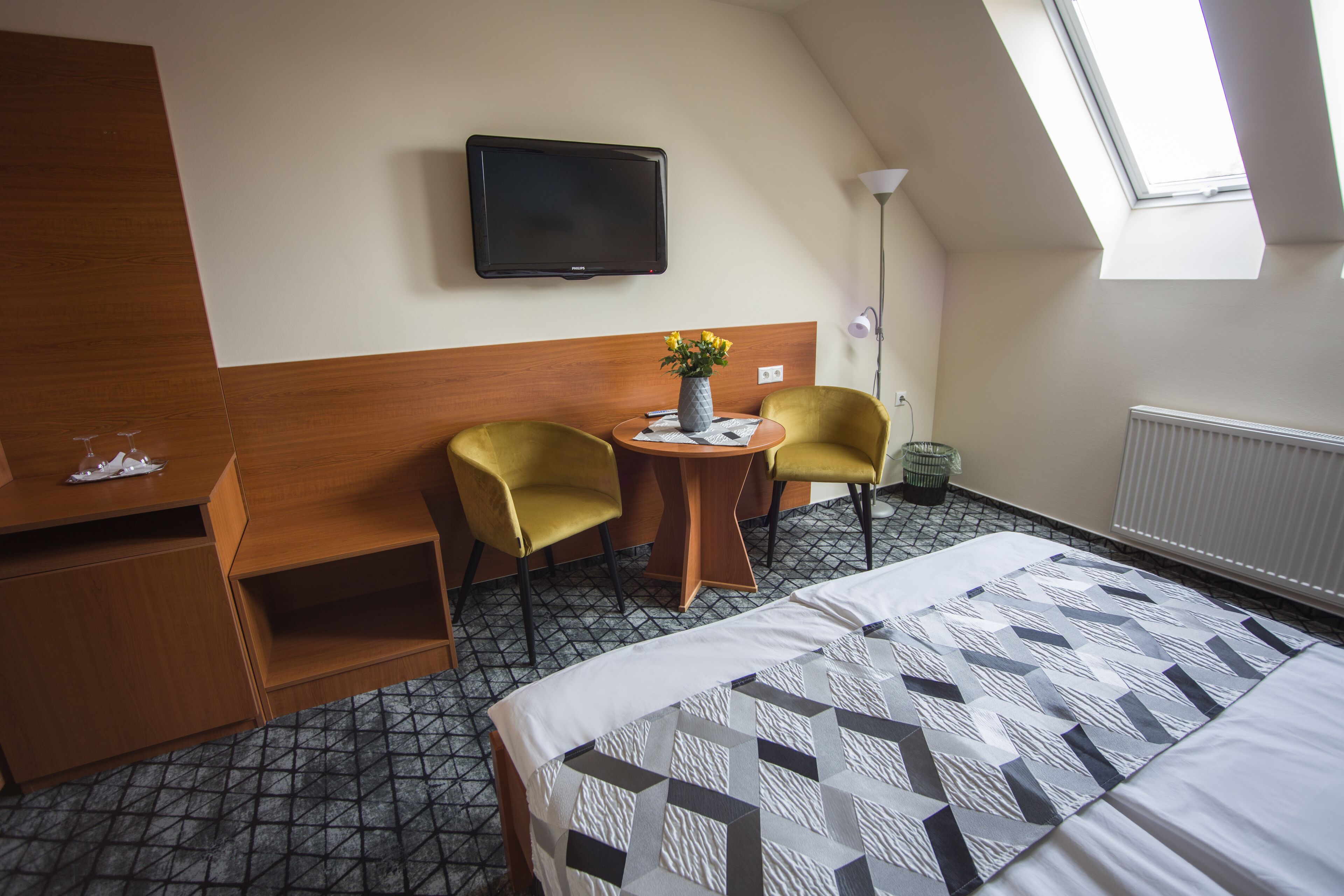 A hotel room with a bed, two yellow chairs, a small table with a vase, a flat-screen TV, and a radiator.