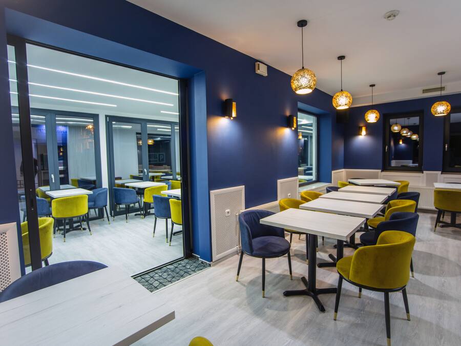 Uma sala de jantar moderna com mesas e cadeiras, cozinha visível e uma grande janela que permite entrada de luz natural.