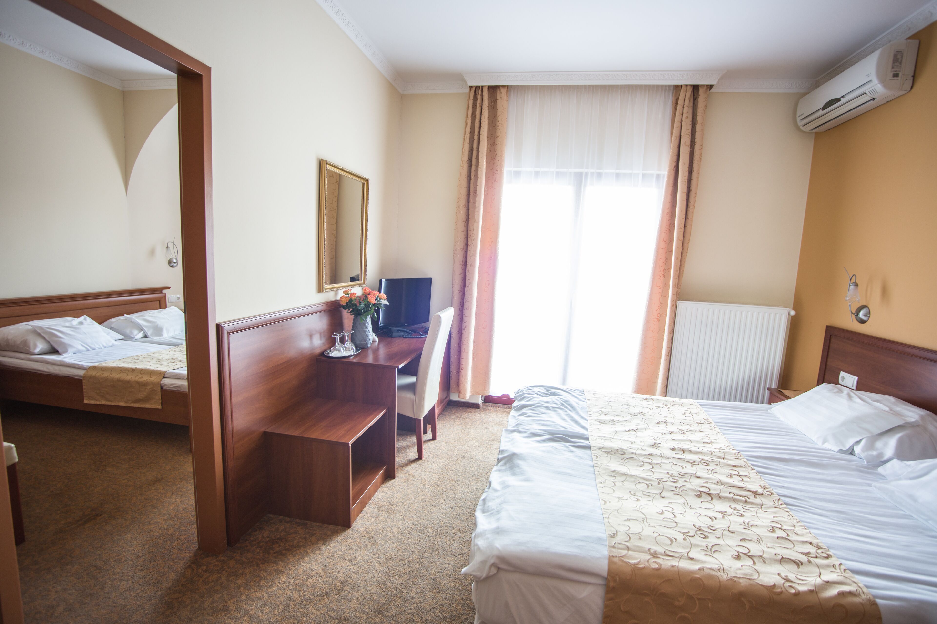 A hotel room with two beds, a desk, a chair, a mirror, and an air conditioning unit.