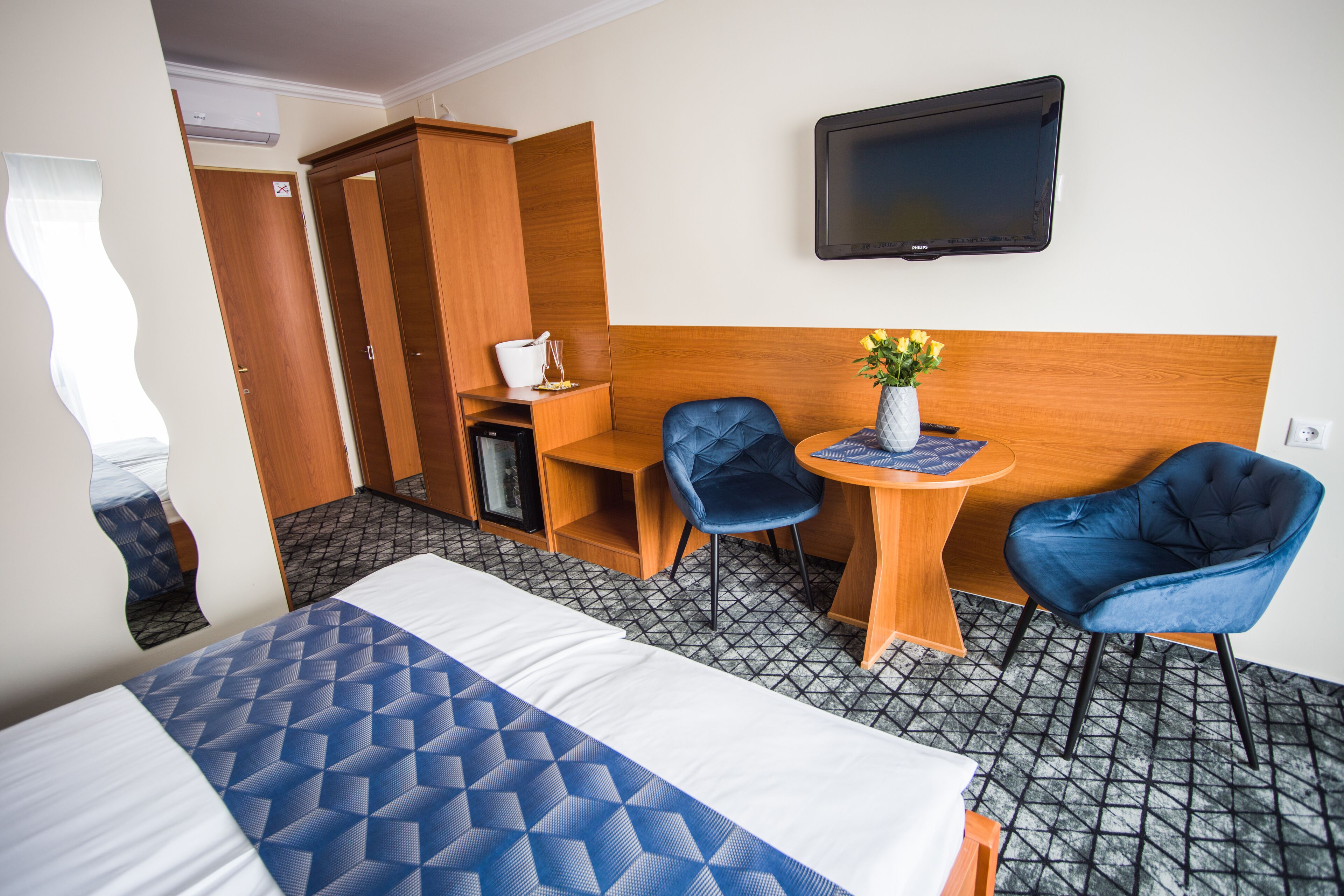 A hotel room with a bed, two blue chairs, a small table with a vase, a TV on the wall, and a wardrobe.