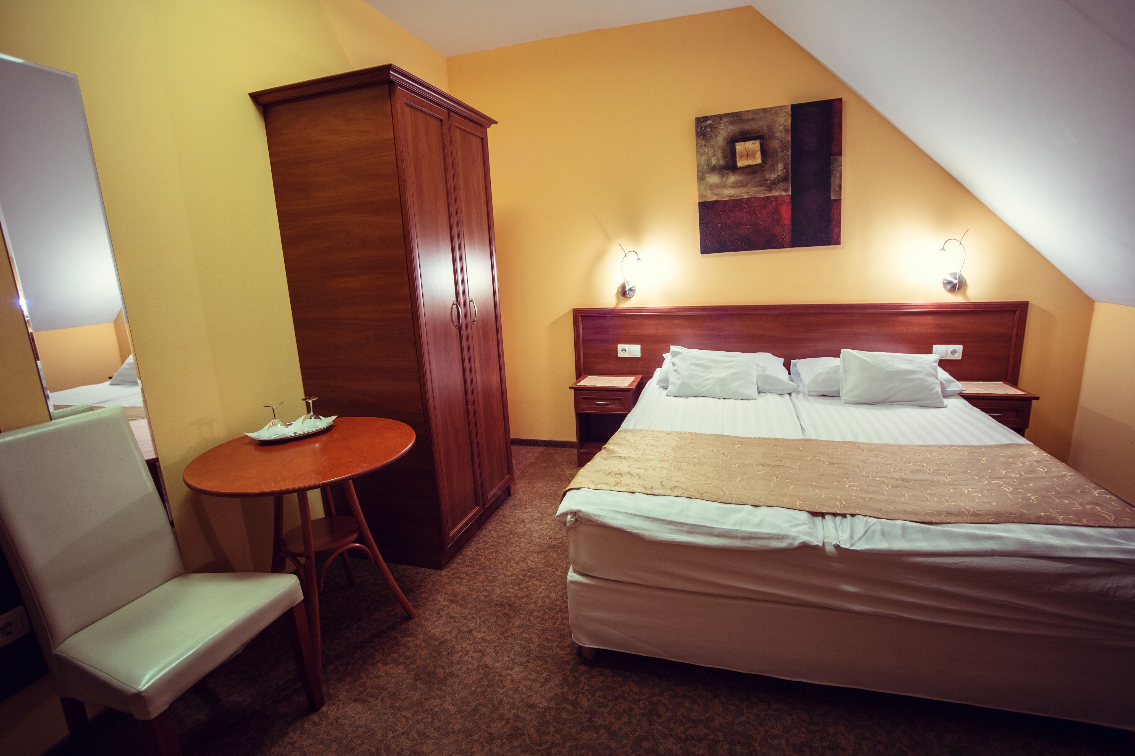 A hotel room with a bed, a wooden wardrobe, a small table with a chair, and a wall-mounted light fixture.
