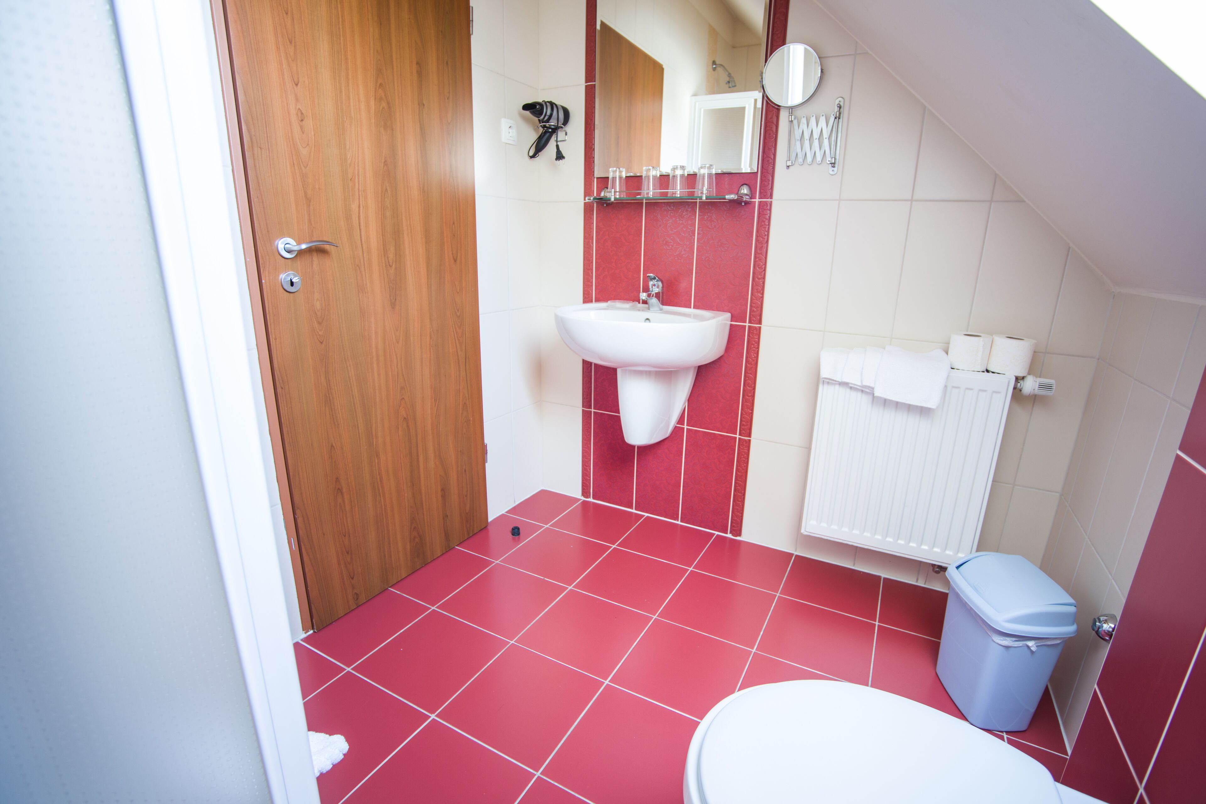 Une petite salle de bain avec une porte en bois, un lavabo blanc, des carreaux rouges, des toilettes blanches et une poubelle.