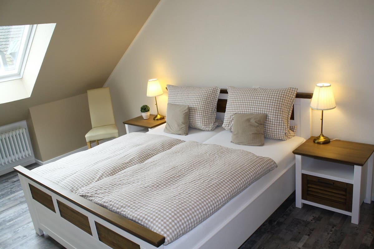 A bedroom with a sloped ceiling, a bed with checkered bedding, two bedside tables with lamps, a chair, and a radiator.