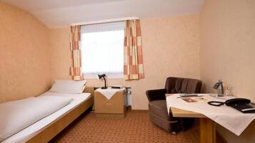 A small hotel room with a single bed, a desk with a chair, a desk lamp, a telephone, and a window with curtains.