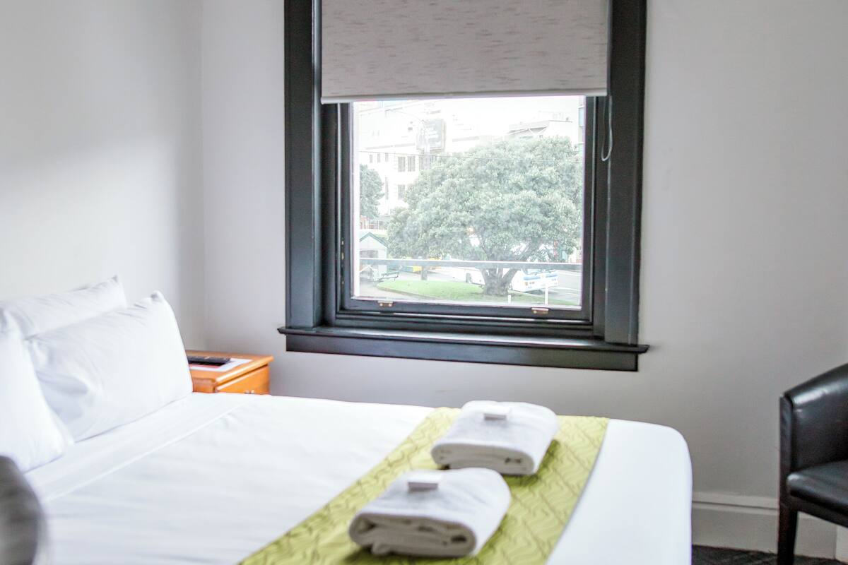 Una habitación de hotel con cama, ventana con vistas a edificios y árboles, una silla y una mesita de noche con lámpara.