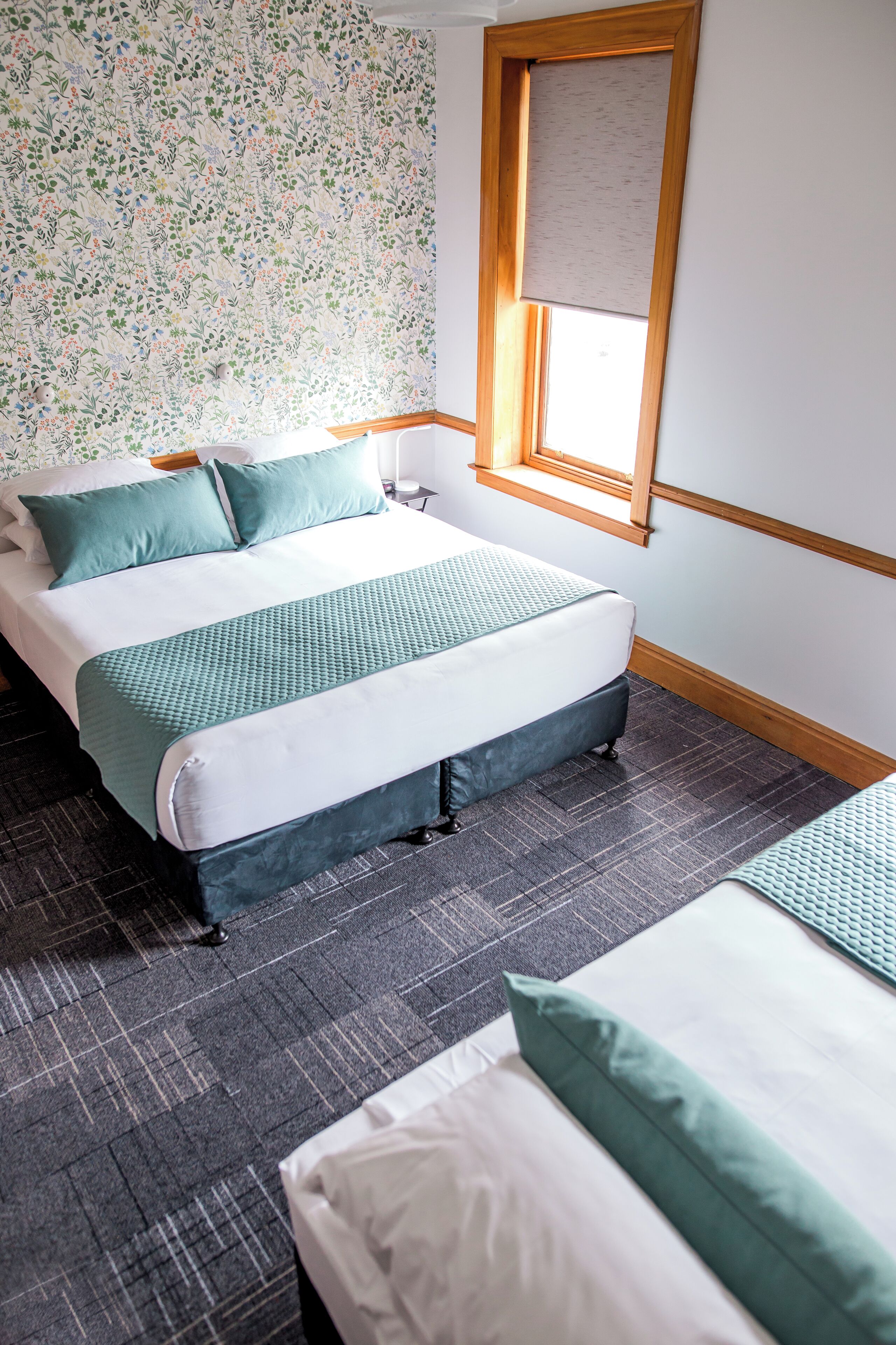 A hotel room with a large bed, a smaller bed, a window with a wooden frame, and a floral wallpaper pattern.