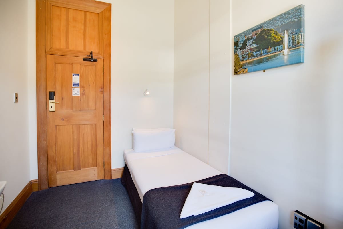 Una habitación pequeña y ordenada con una puerta de madera, una cama individual con colchón blanco y ropa de cama oscura, y un cuadro de un paisaje urbano colgado en la pared.