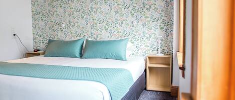 Una cama bien hecha con ropa de cama color turquesa, una pared con estampado floral y una mesita de noche de madera con una lámpara.