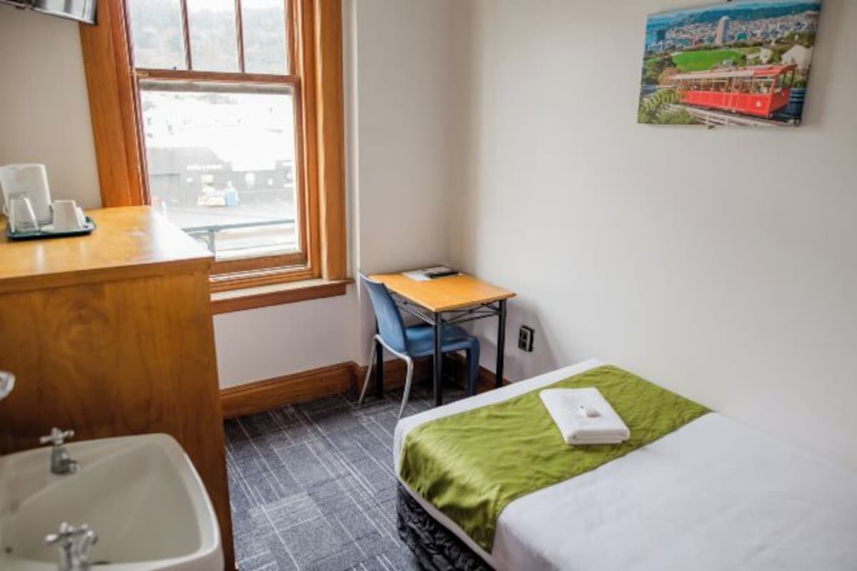 A small hotel room with a bed, a desk, a chair, a window, and a sink.