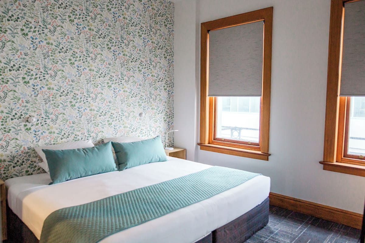Una cama bien hecha con una colcha verde y almohadas, un estampado floral en el papel tapiz y una ventana con marco de madera y persianas.
