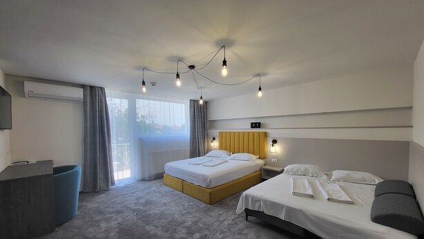 A modern hotel room with a large bed, a smaller bed, a desk, and a chair. There is a large window with curtains, and the room has a neutral color scheme. - XO Residence (Arad)