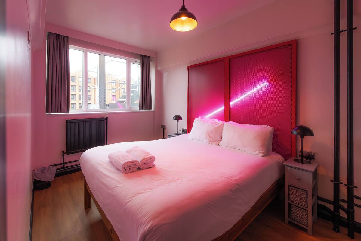 A bedroom with a large bed, a window, a radiator, a nightstand, and a lamp.