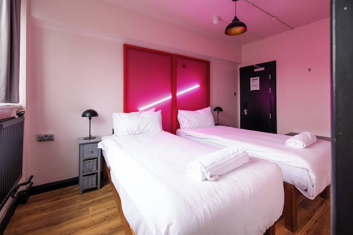 A hotel room with two beds, a red sliding door, a black door, a small desk with a lamp, and a wooden floor.