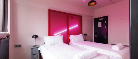 A hotel room with two beds, a red sliding door, a black door, a small desk with a lamp, and a wooden floor.