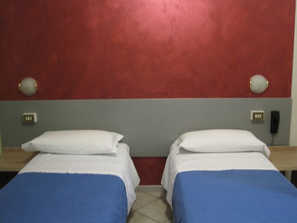 Quarto de hotel com duas camas de solteiro, cada uma com colcha azul e almofadas brancas. Há mesas de cabeceira com abajures e um telefone na parede.