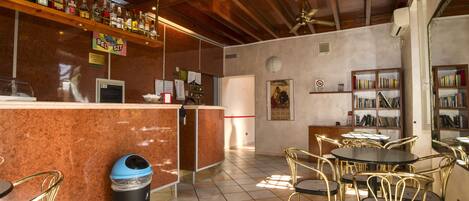 Un espace bar comprenant un comptoir, des étagères remplies de bouteilles, une poubelle et un coin salon avec des chaises et une table.