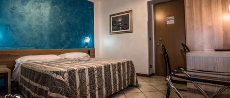 A hotel room with a bed, a nightstand, a wall clock, a framed picture, a door with a sign, and a chair.