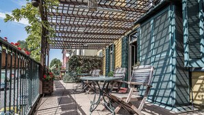 Ένας εξωτερικός χώρος με τραπέζι και καρέκλες, μια πέργκολα και ένα κτίριο με παντζούρια.