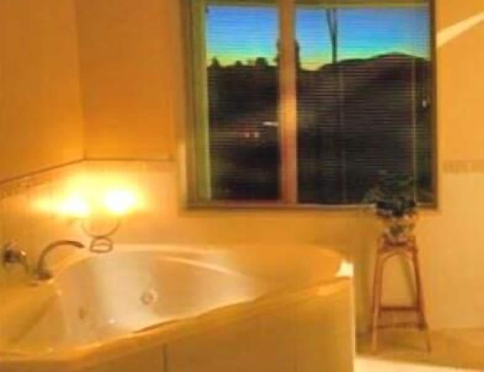 A bathroom with a large bathtub, a window with blinds, and a small stool.