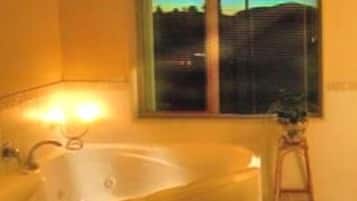 A bathroom with a large bathtub, a window with blinds, and a small stool.