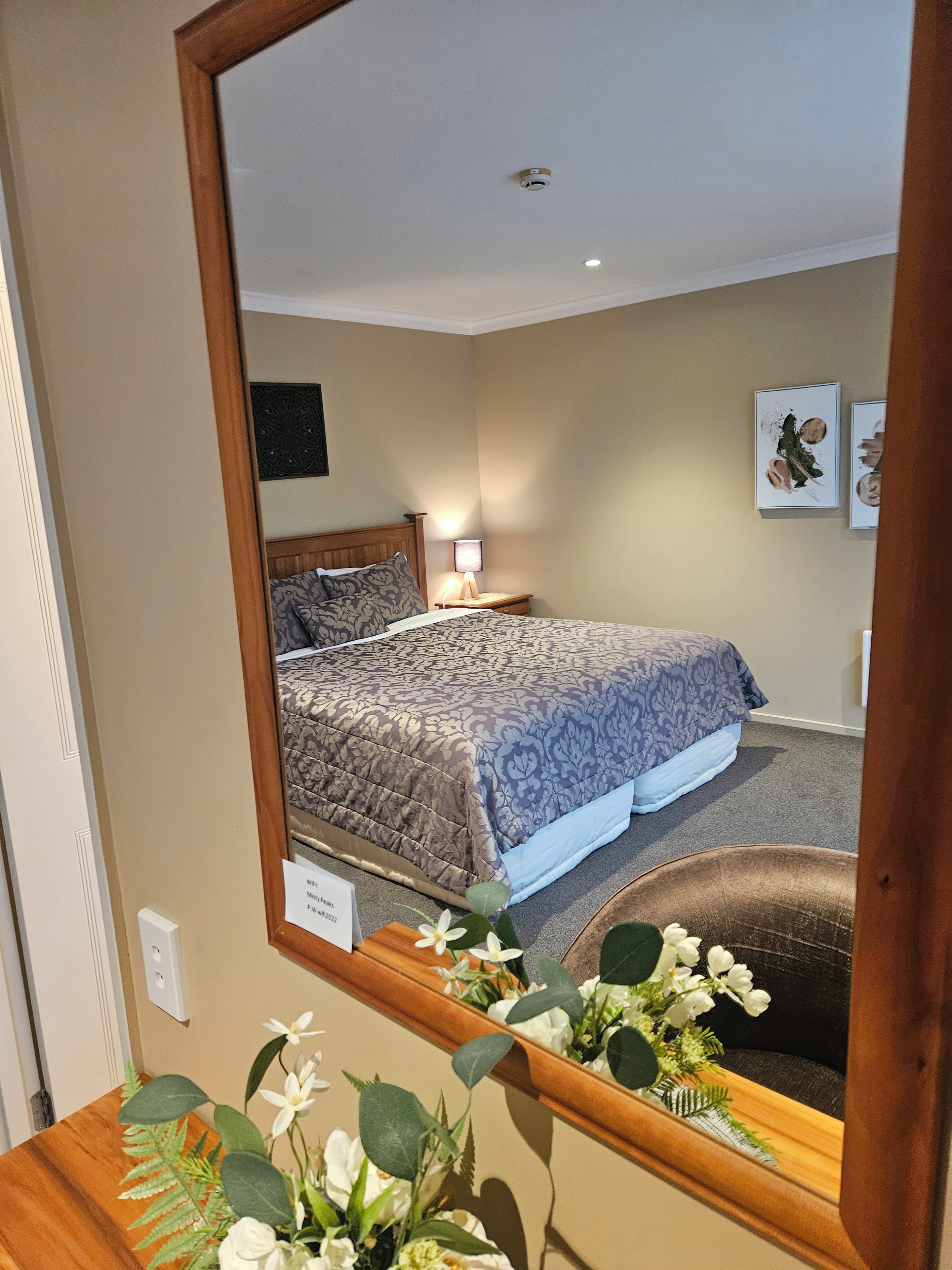 A bedroom with a bed, a nightstand, a wall-mounted lamp, and a mirror reflecting the room.