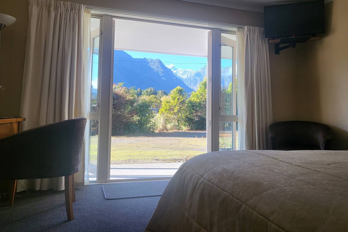 Quarto de hotel com cama, uma cadeira e televisão. O quarto possui uma janela grande com cortinas, oferecendo vista para montanhas e vegetação.