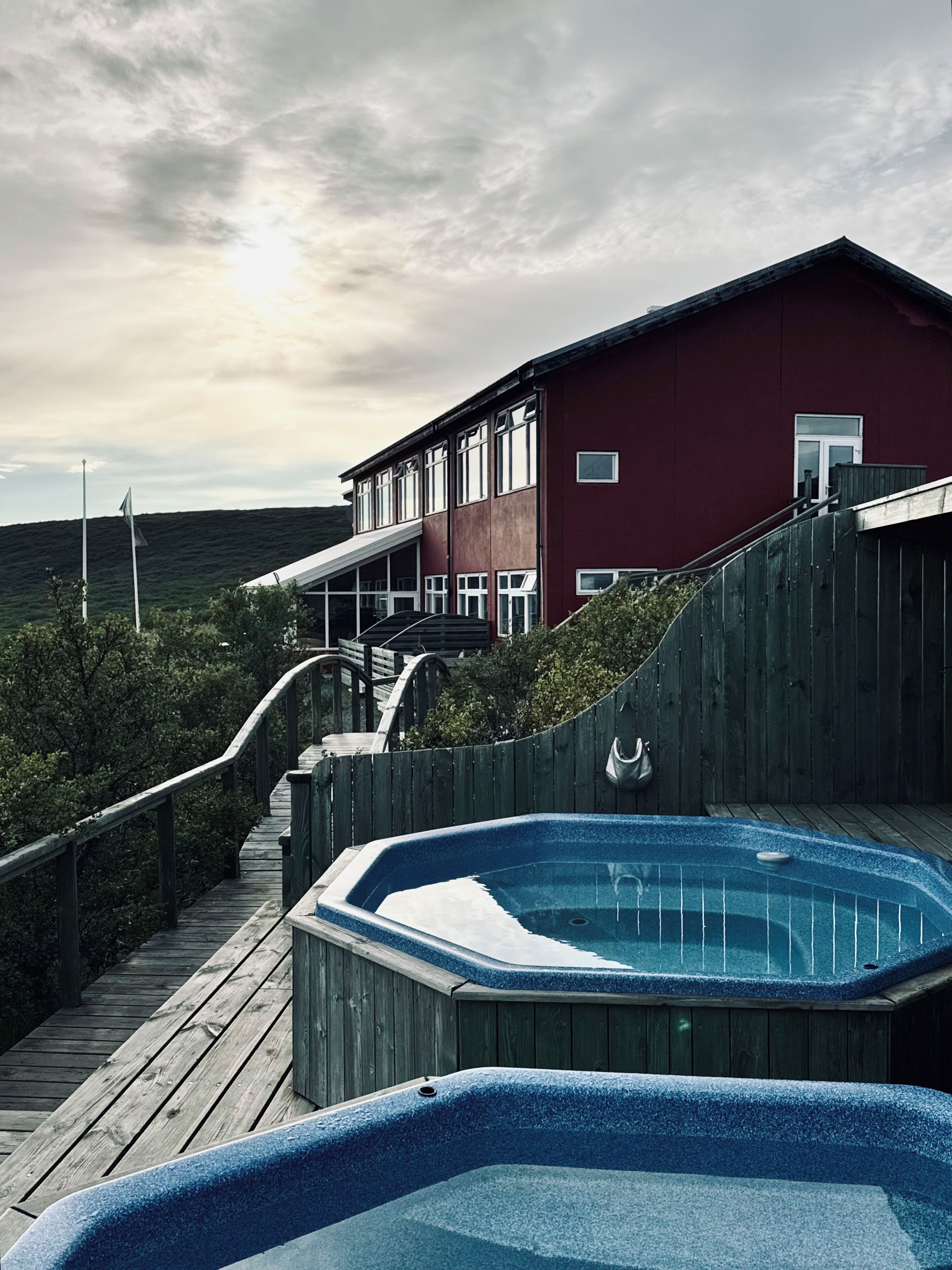 A wooden deck leading to a hot tub with a view of a red building and a cloudy sky.
