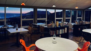 A spacious room with large windows, a round table with orange chairs, and a bookshelf filled with books. - Hotel Glymur (Hvalfjarðarsveit)
