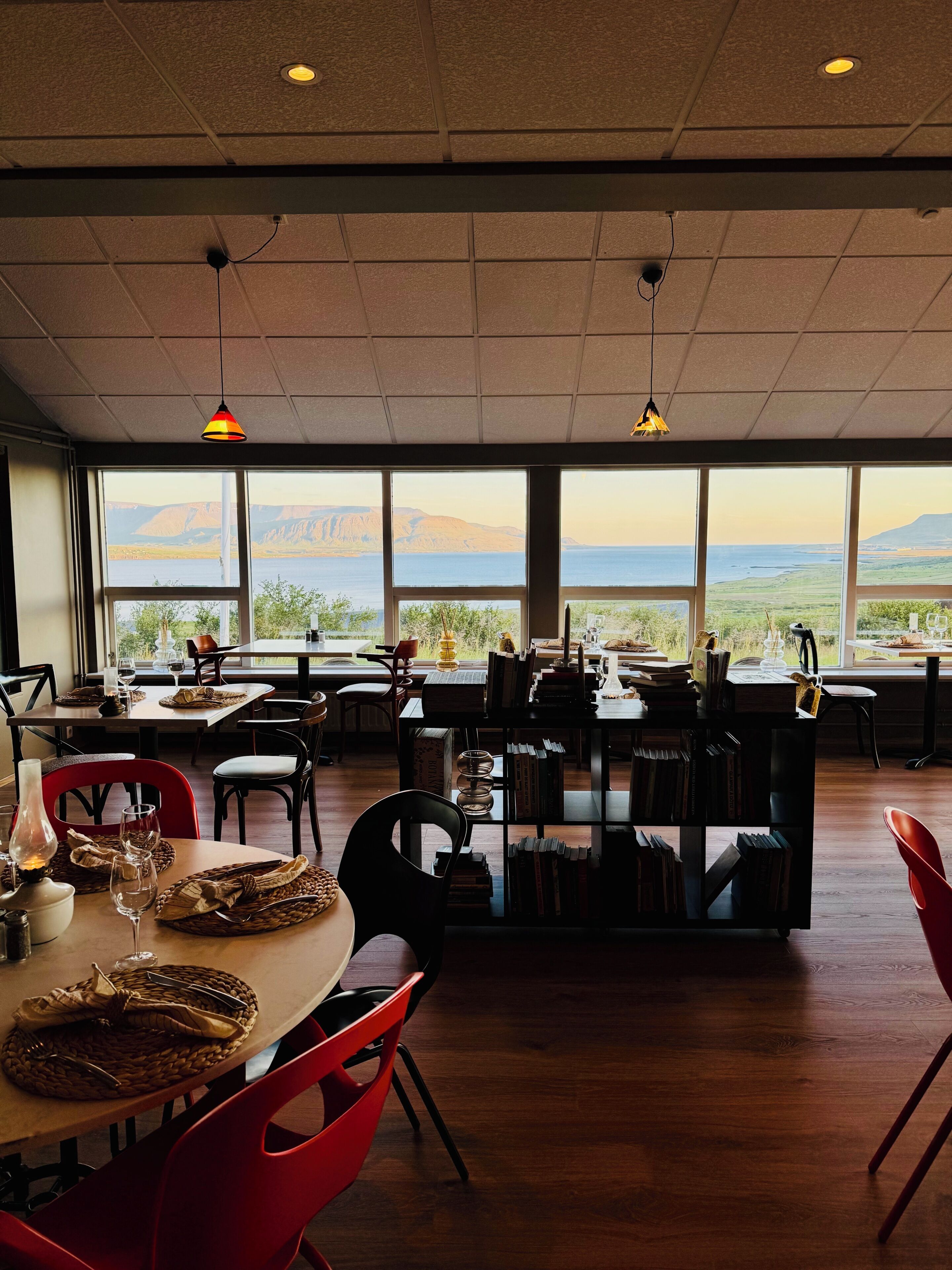 Comedor con vistas a montañas y un lago, con mesas de madera, sillas rojas e iluminación colgante.