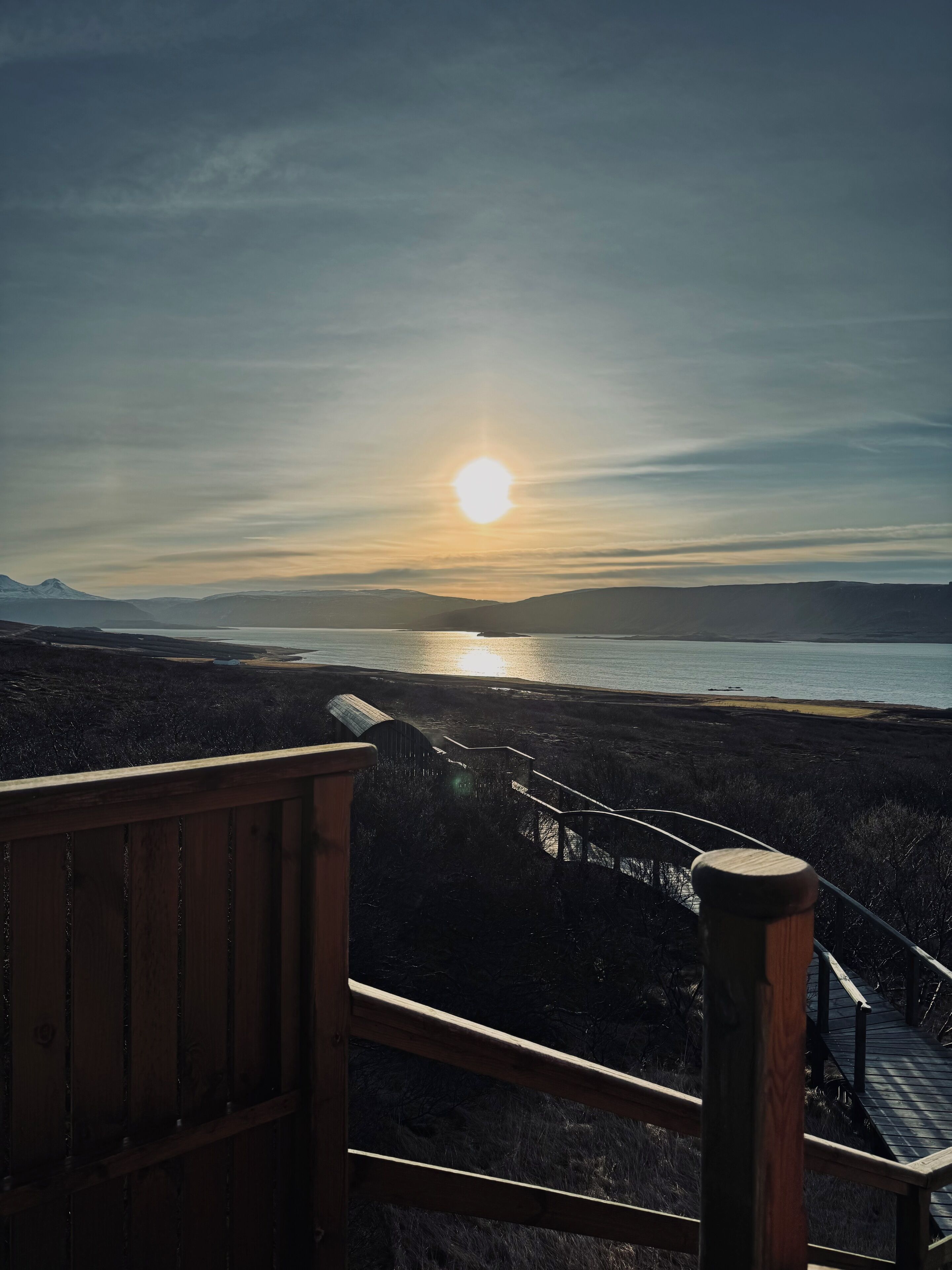 A wooden staircase leading to a viewing platform overlooking a body of water and distant mountains during sunset.