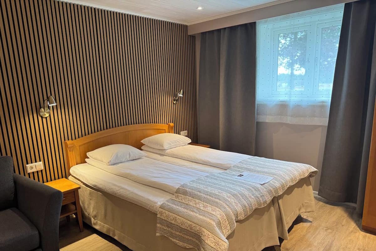 Ein Doppelbett mit Holzfußteil, ein Nachttisch, eine Lampe und ein Fenster mit Vorhängen.