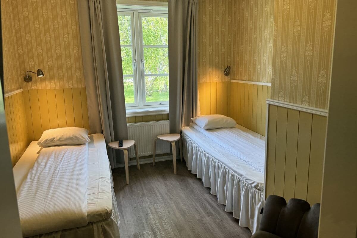 Ein Zimmer mit zwei Einzelbetten, einem kleinen Tisch und einem Fenster mit Vorhängen.
