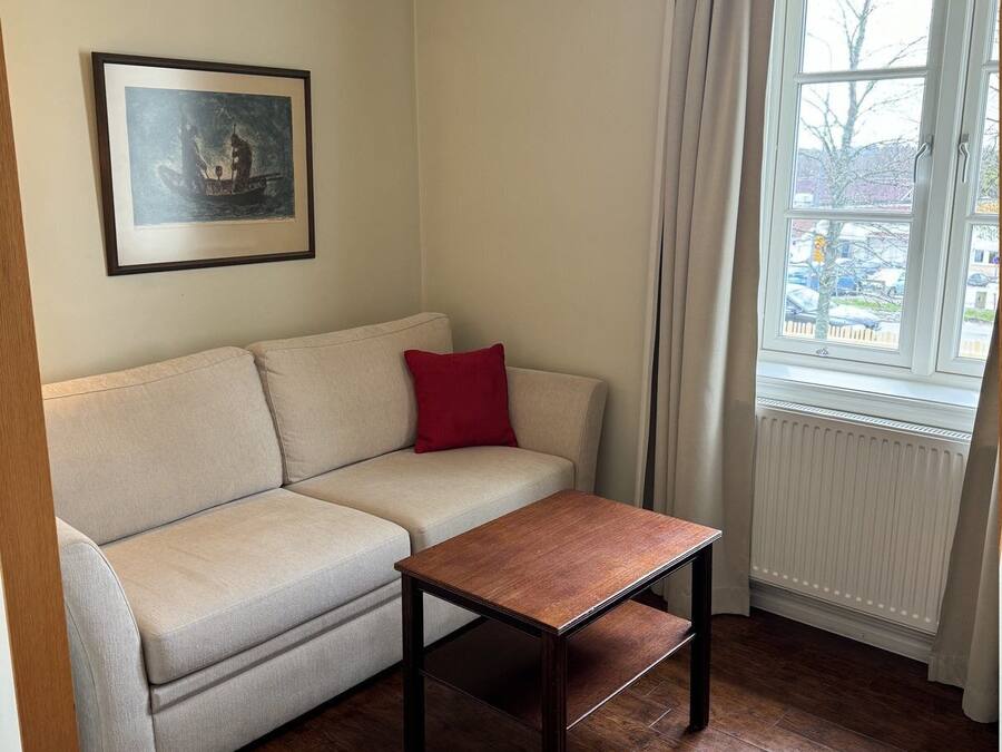A room with a beige sofa, a small wooden table, a framed picture on the wall, and a window with curtains.