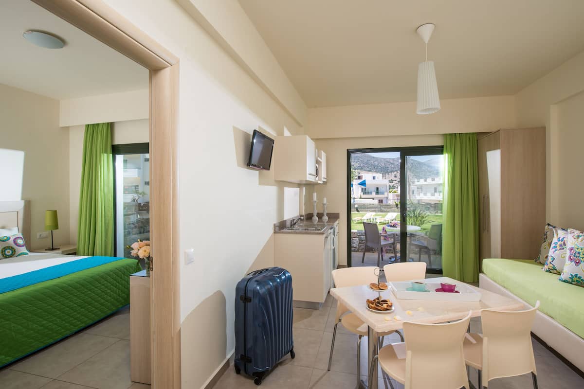 A hotel room with a bed, a desk with a chair, a table with a chair, a suitcase, a TV mounted on the wall, and a balcony with a view of the outside.