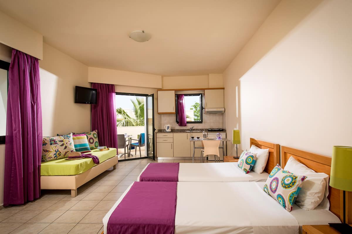 A hotel room with two beds, a sofa, a kitchenette, and a balcony with curtains.