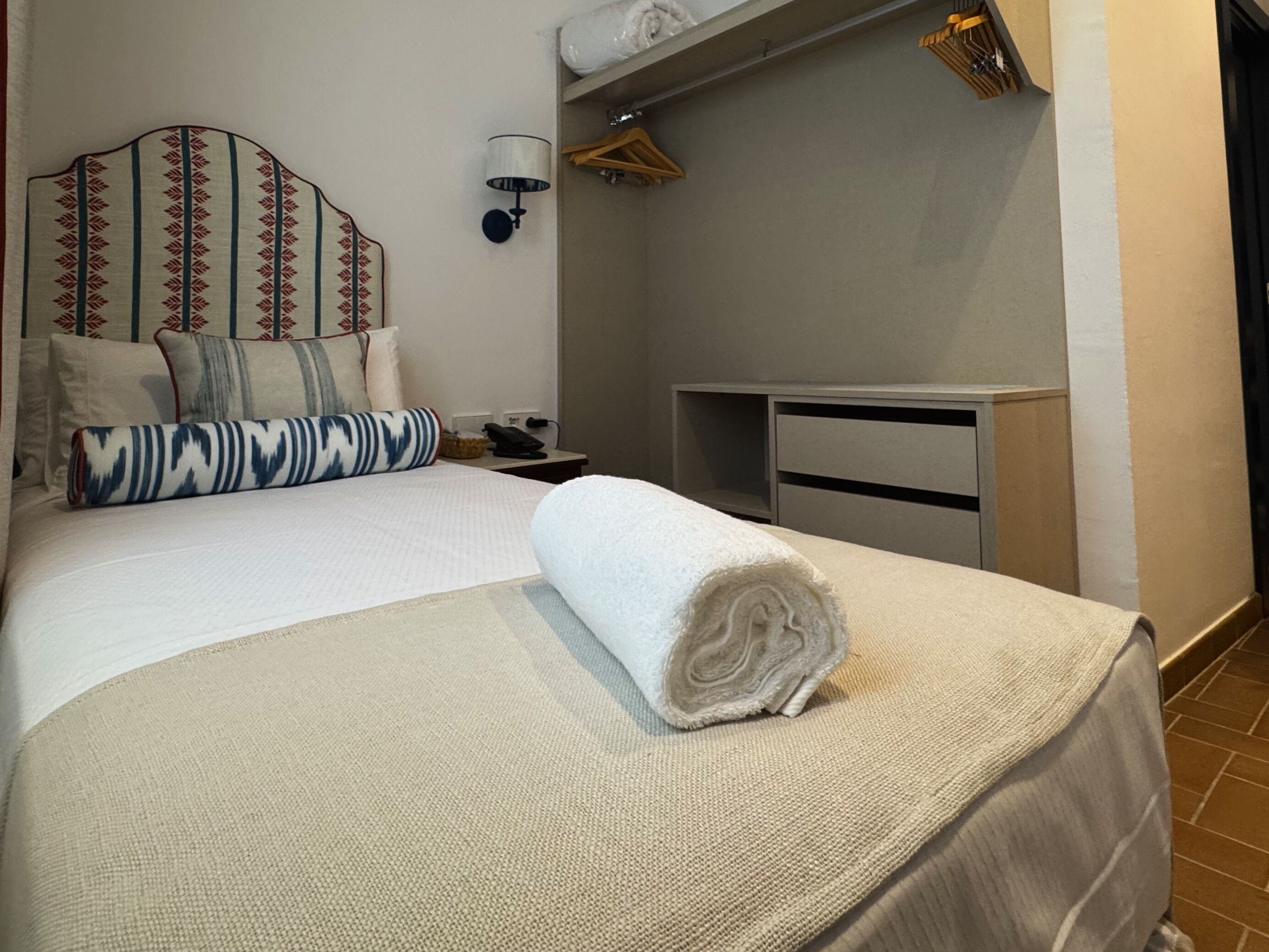 A neatly made bed with a rolled towel, a bedside table with a lamp, and a wardrobe.