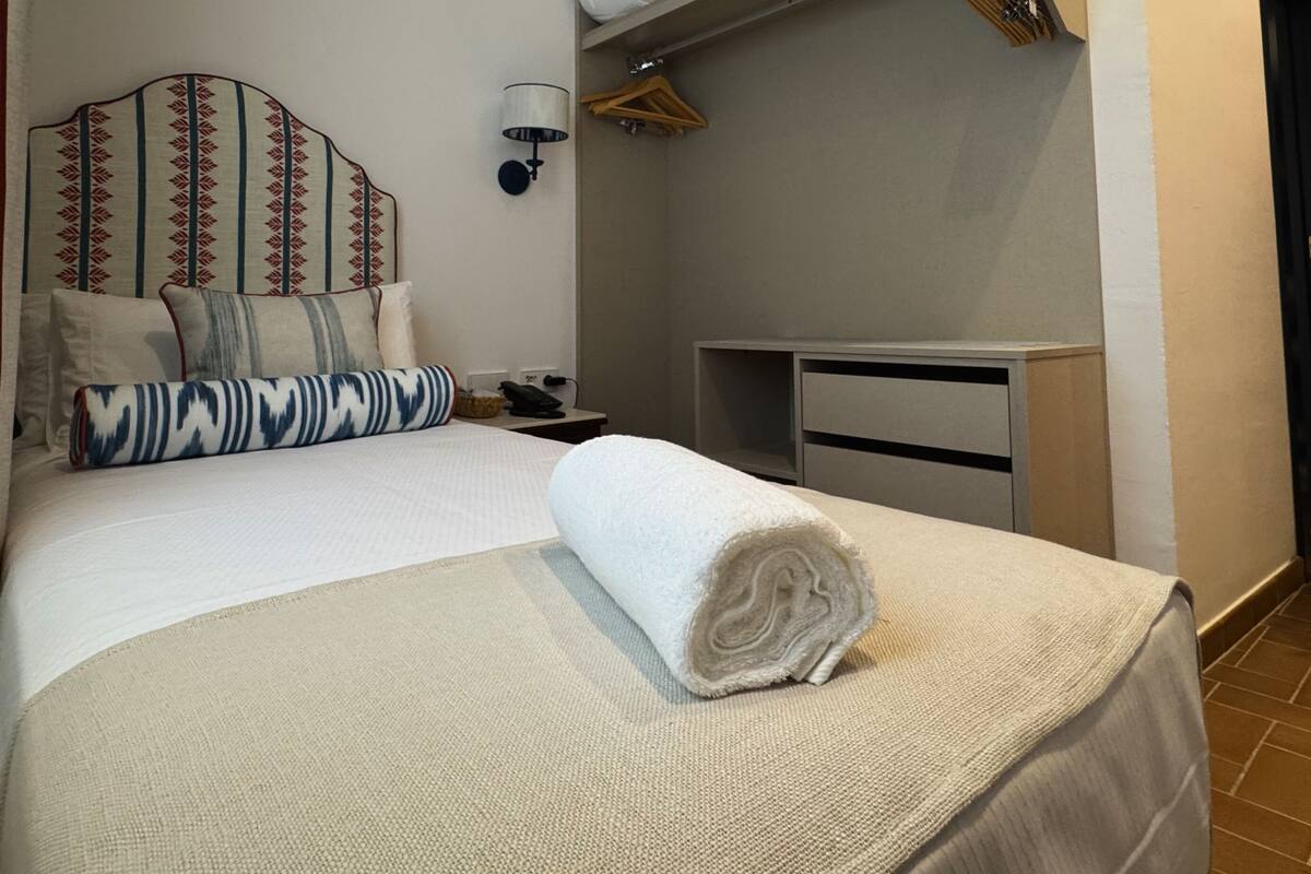 A neatly made bed with a rolled towel, a bedside table with a lamp, and a wardrobe.