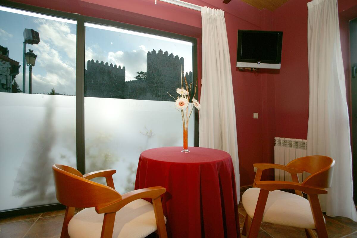 Una habitación con un ventanal grande, una mesa con mantel rojo, dos sillas de madera y un televisor instalado en la pared.