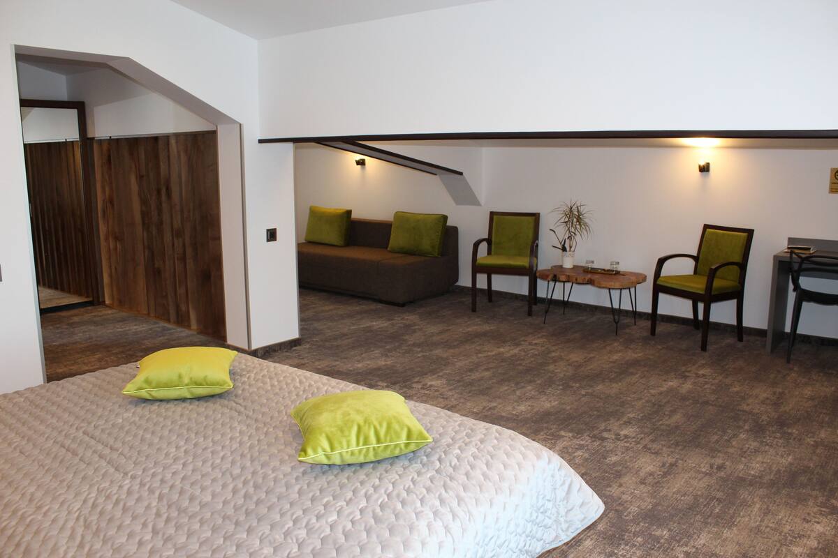 A modern hotel room with a large bed, two green pillows, a sofa, two chairs, a small table, and a desk.