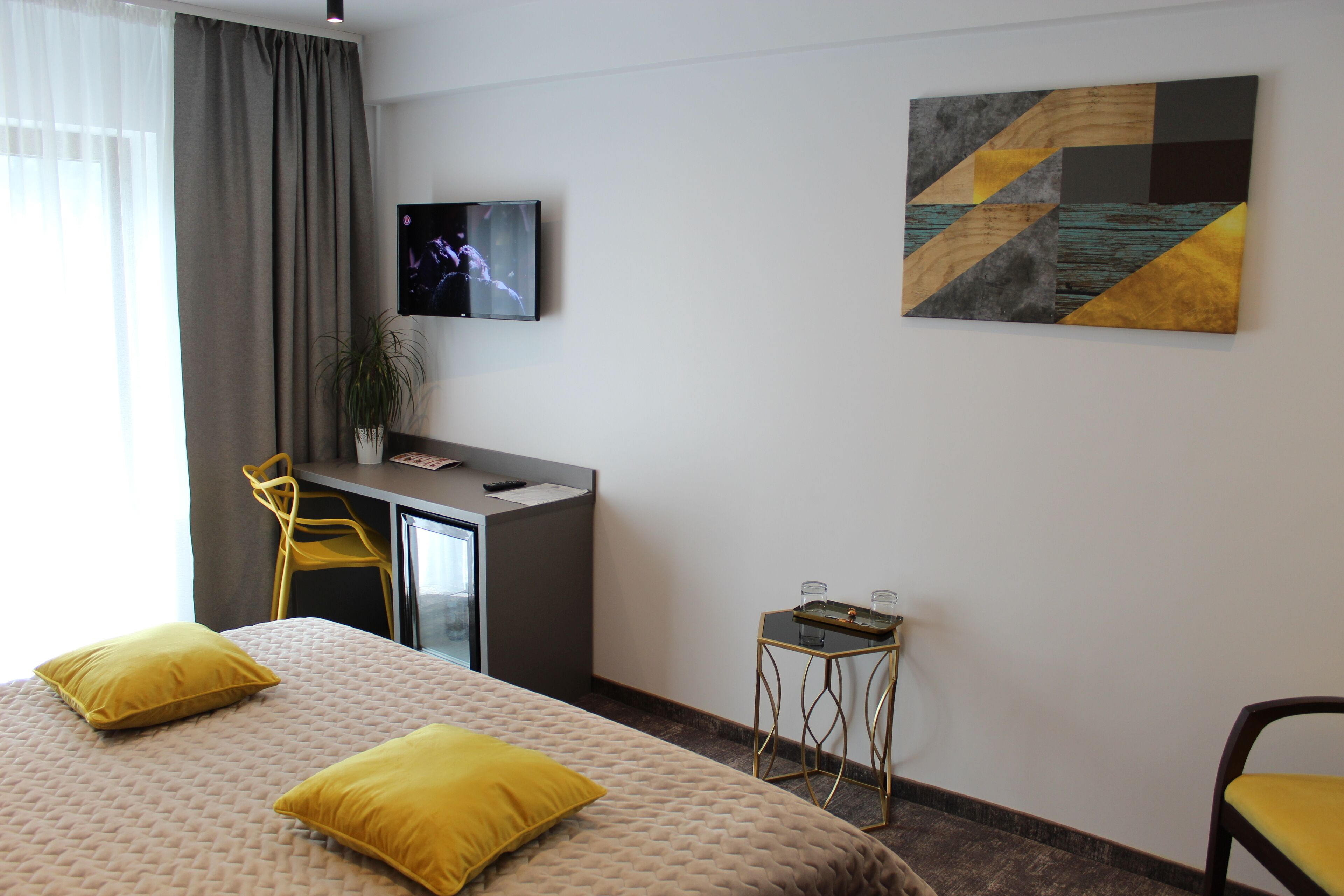 A modern hotel room with a bed, a desk, a TV, and a painting on the wall.