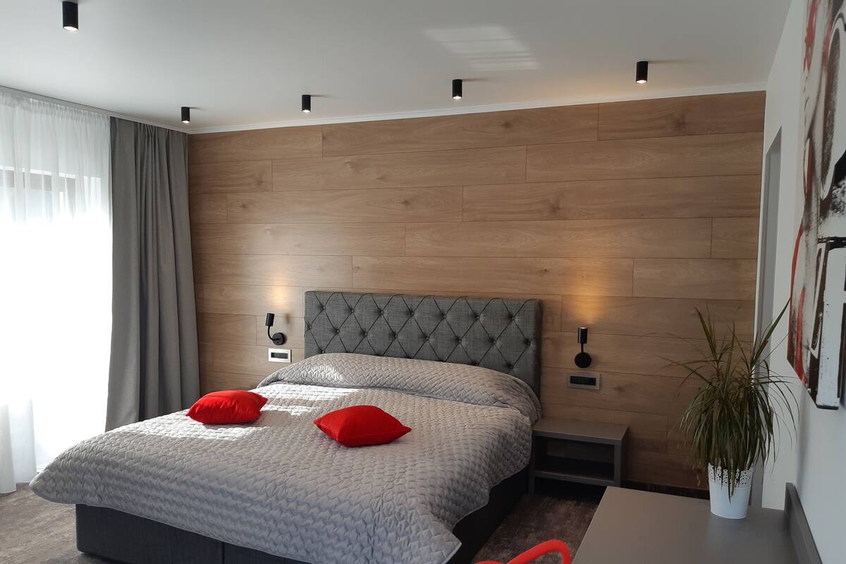 A modern bedroom with a large bed, grey and red pillows, a grey upholstered headboard, bedside tables with lamps, a wooden wall panel, and a potted plant.