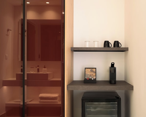 A modern bathroom with a glass shower enclosure, a sink with toiletries, and a shelf with a coffee maker and a picture frame.