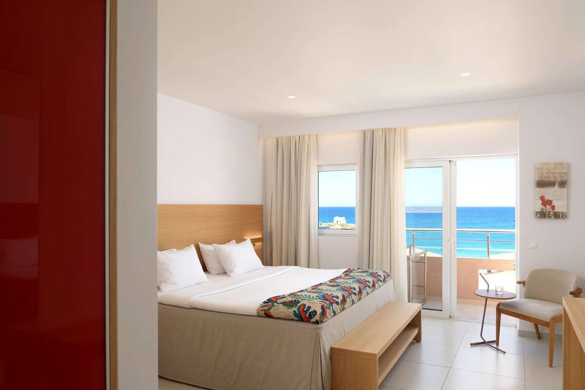 A hotel room with a bed, a bench, a chair, a small table, and a view of the sea through large windows.
