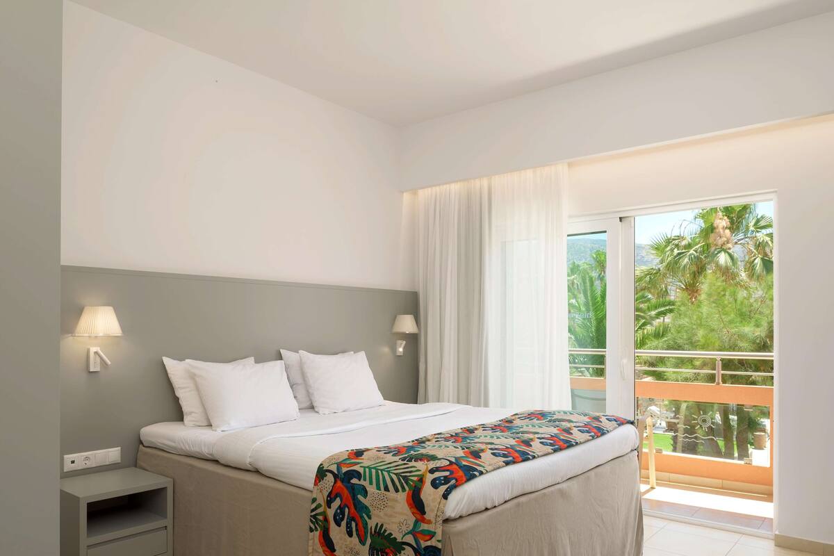 A hotel room with a bed, bedside lamps, a nightstand, and a view of the outdoors.